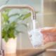 A man filling a glass with tap water from a kitchen sink with a silver faucet and a potted plant in the background.