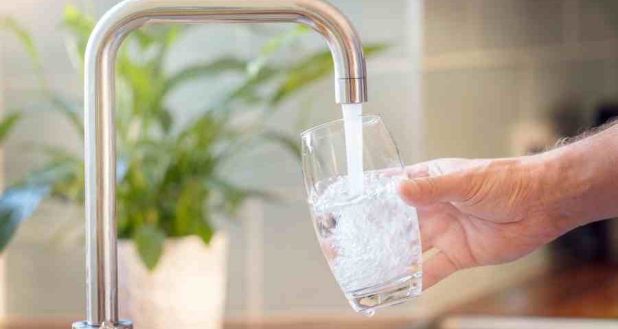 A man filling a glass with tap water from a kitchen sink with a silver faucet and a potted plant in the background.