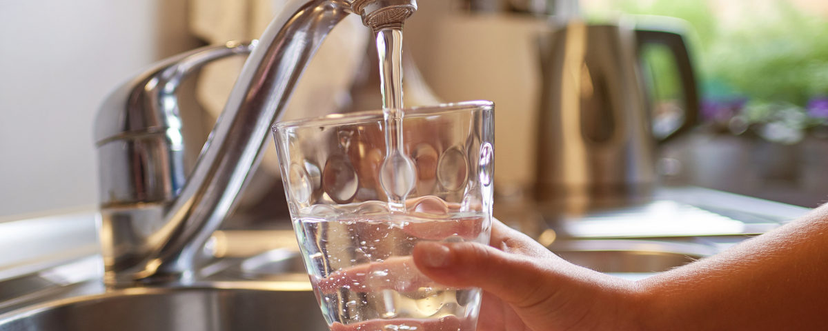 A hand fills a short glass with running water from a silver faucet above the sink in a kitchen.