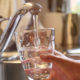 A hand fills a short glass with running water from a silver faucet above the sink in a kitchen.