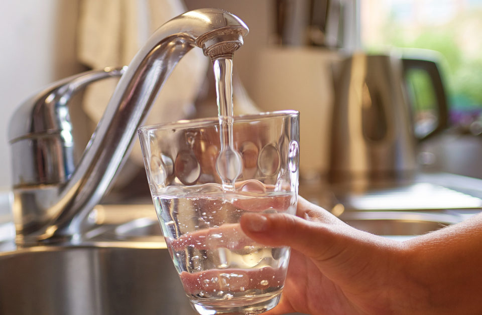 A hand fills a short glass with running water from a silver faucet above the sink in a kitchen.