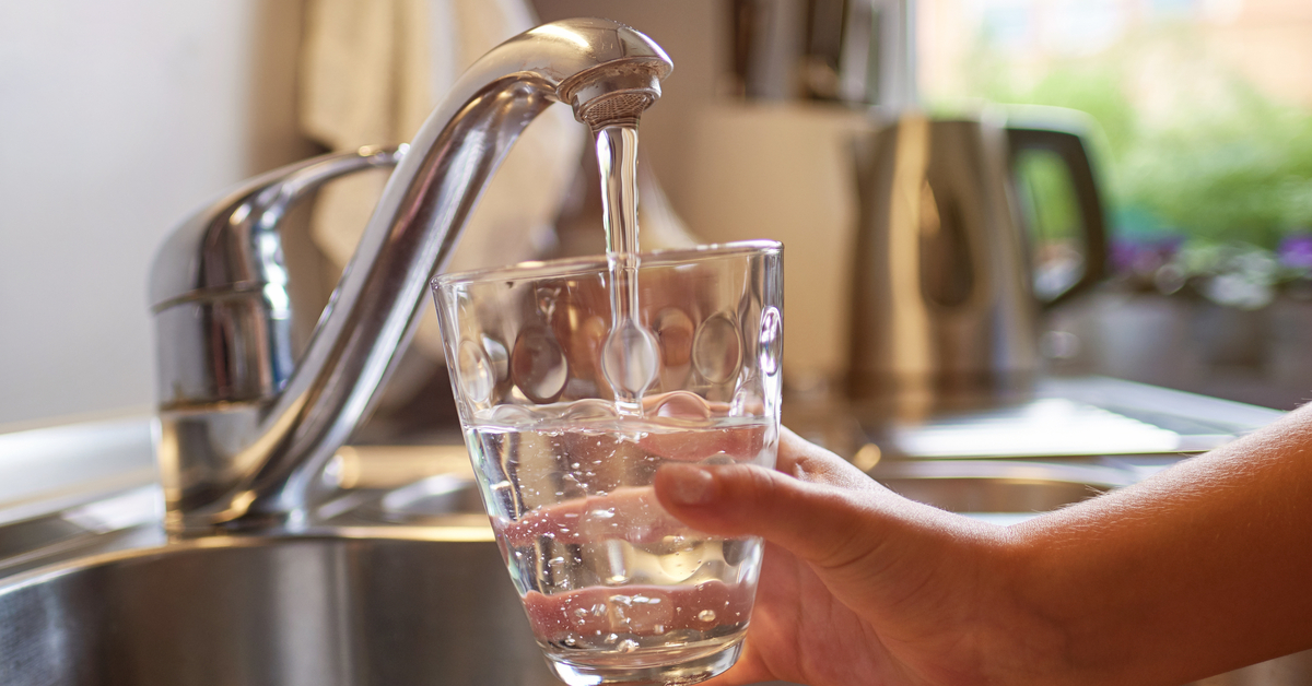 A hand fills a short glass with running water from a silver faucet above the sink in a kitchen.