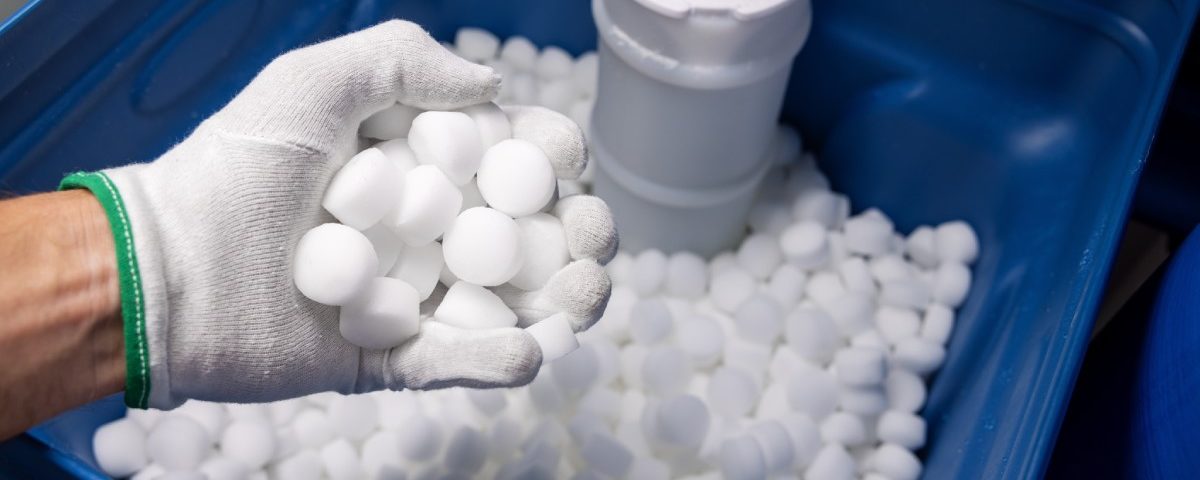 An open water softener tank filled with white salt tablets. A man's hand wearing a glove holds a handful of tablets.