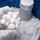 An open water softener tank filled with white salt tablets. A man's hand wearing a glove holds a handful of tablets.