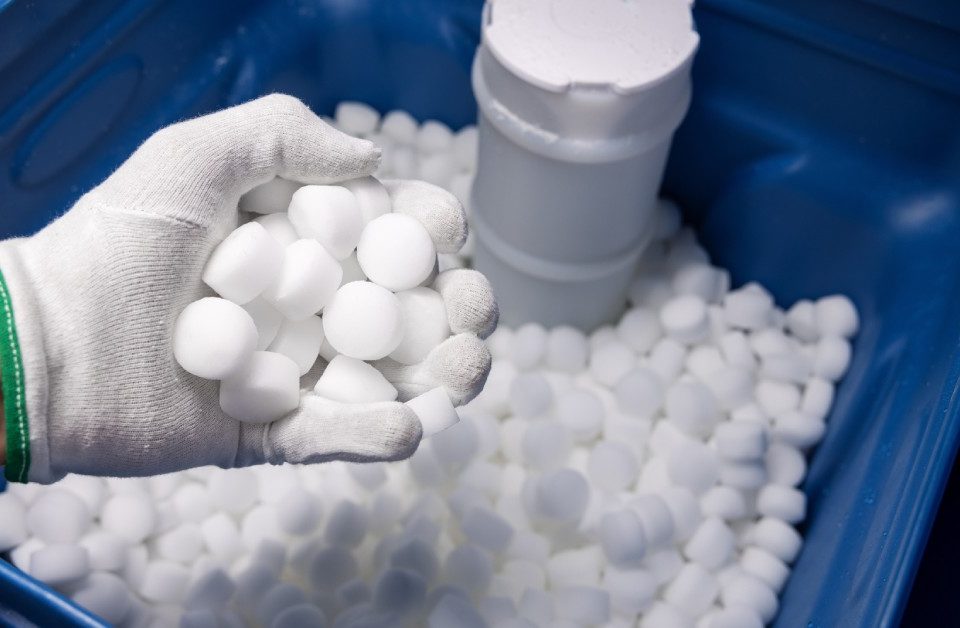 An open water softener tank filled with white salt tablets. A man's hand wearing a glove holds a handful of tablets.