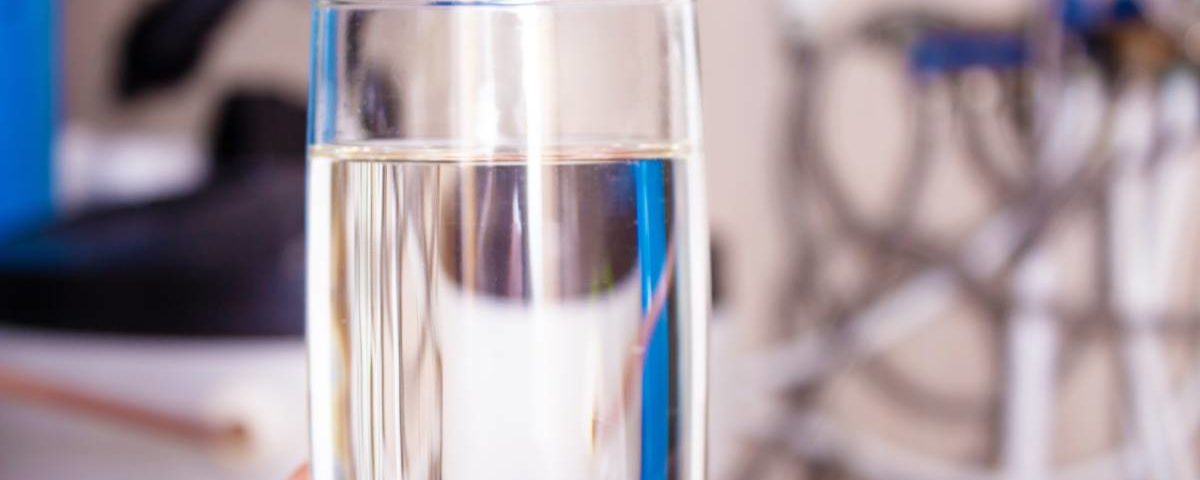 A person's hand is holding a full tall glass of water in the foreground. A blurry water softener is in the background.