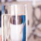 A person's hand is holding a full tall glass of water in the foreground. A blurry water softener is in the background.