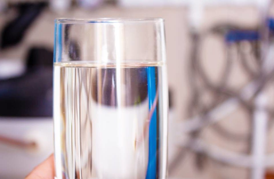 A person's hand is holding a full tall glass of water in the foreground. A blurry water softener is in the background.