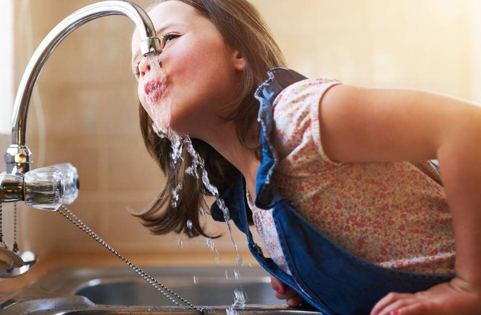 A little girl with brown hair in denim dungarees climbing onto the kitchen counter to drink directly from the sink faucet.