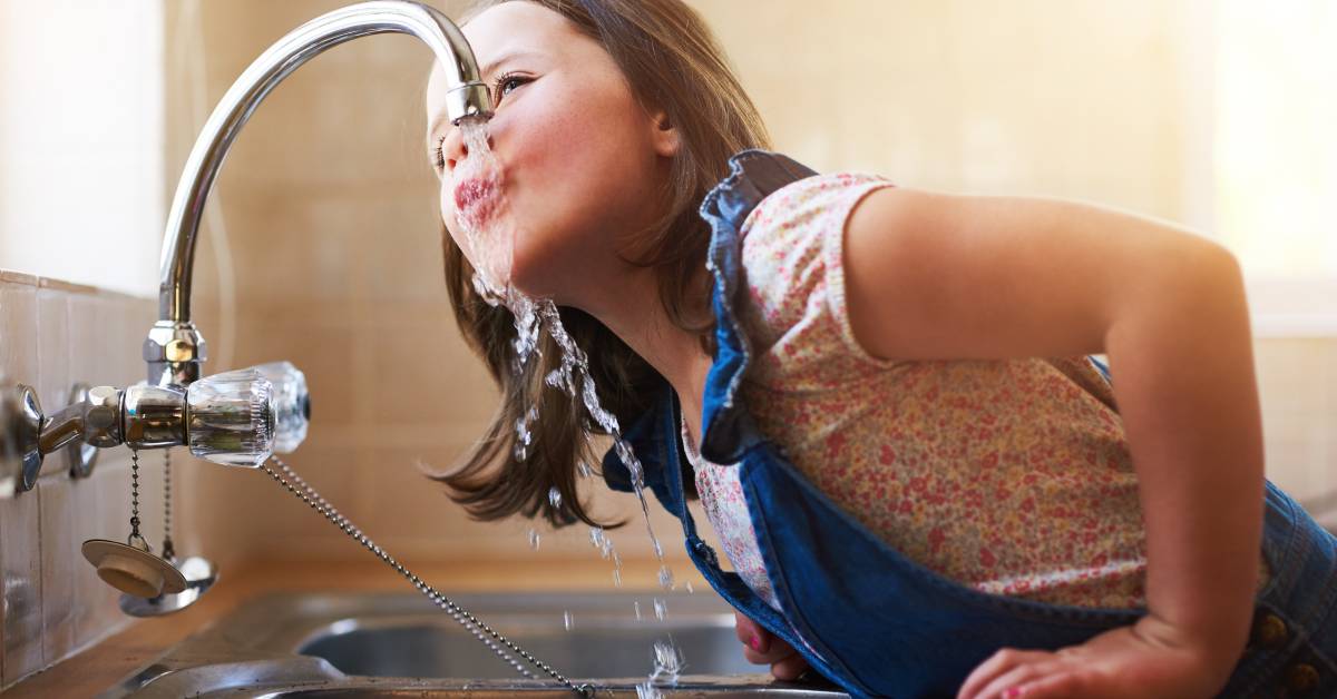 A little girl with brown hair in denim dungarees climbing onto the kitchen counter to drink directly from the sink faucet.