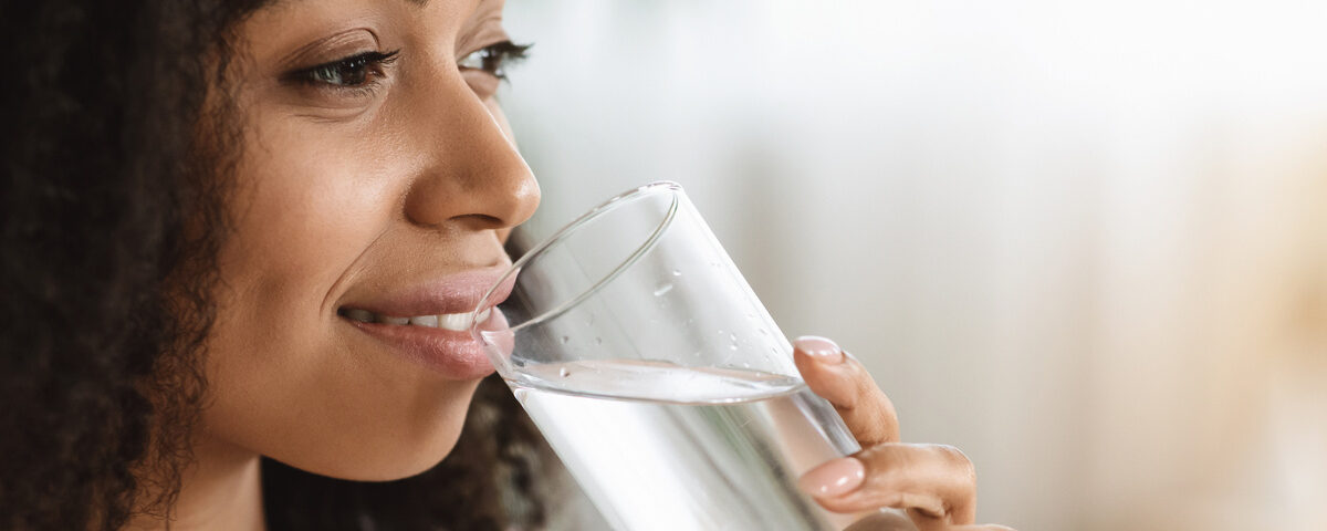 A smiling black woman with textured hair staring off into the distance while enjoying a clear glass of water.