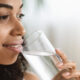 A smiling black woman with textured hair staring off into the distance while enjoying a clear glass of water.