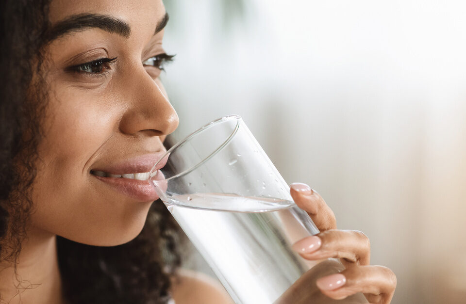 A smiling black woman with textured hair staring off into the distance while enjoying a clear glass of water.