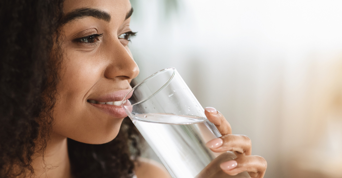 A smiling black woman with textured hair staring off into the distance while enjoying a clear glass of water.