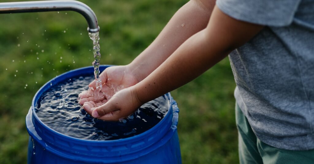 A little boy in shorts washes his hands at an outdoor pump. A large blue bucket catches the excess water.