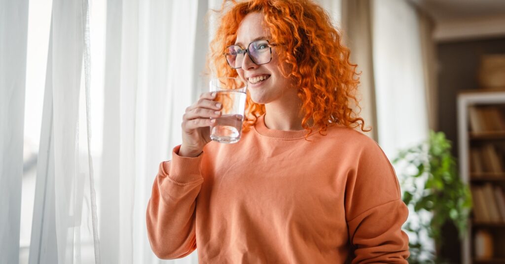 A smiling woman in a sweatshirt holds a glass of water. She's standing next to a window, and there's a bookcase behind her.