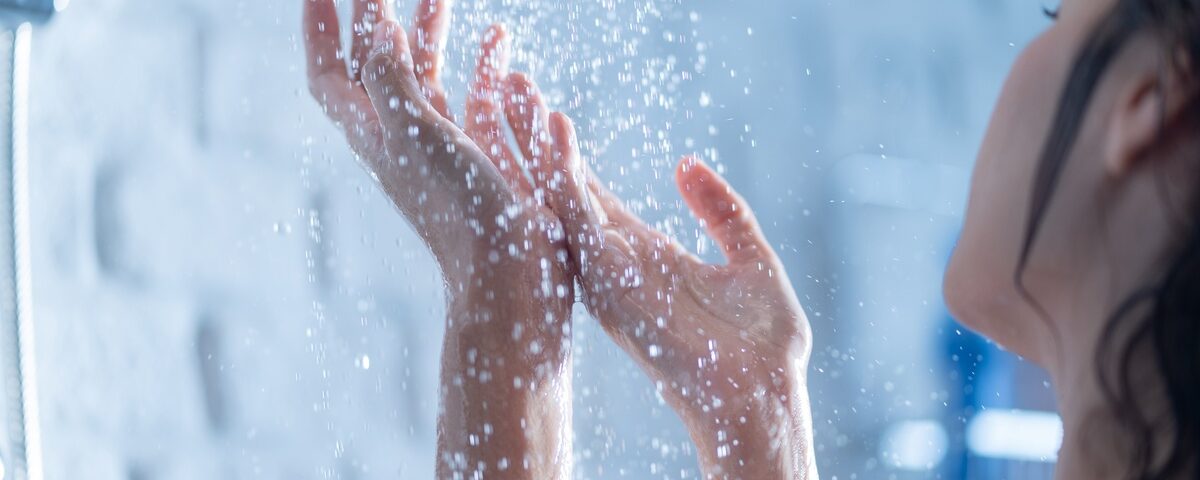 A woman standing inside a shower watches water pour onto her hands. She's in front of a white wall with blurred accents.