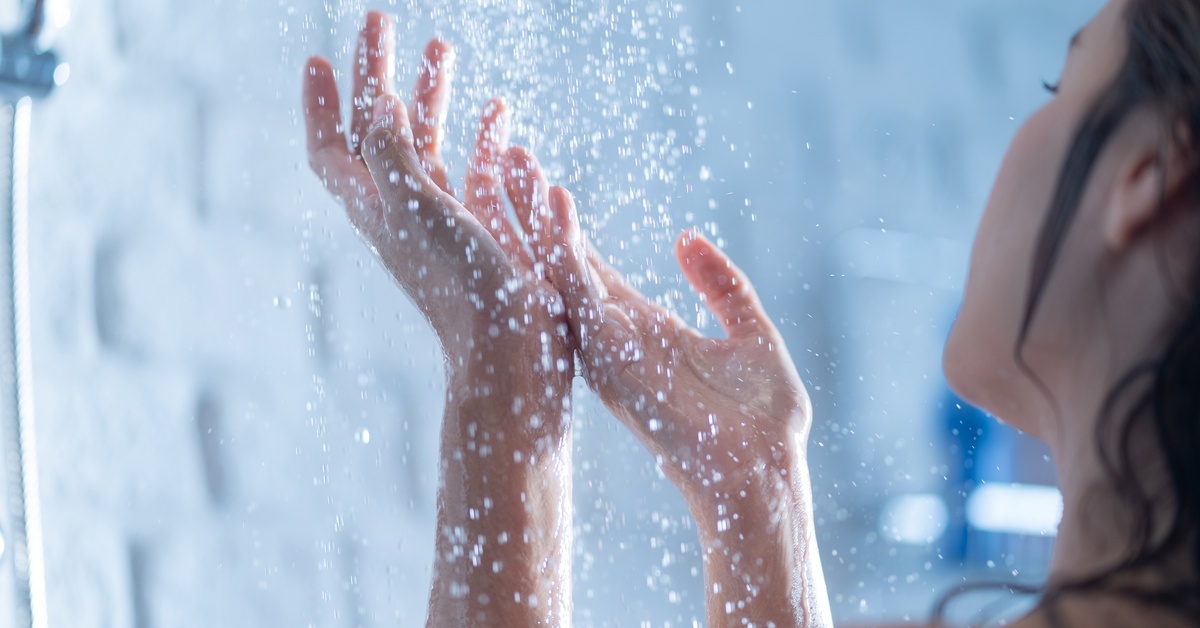 A woman standing inside a shower watches water pour onto her hands. She's in front of a white wall with blurred accents.